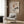 May include: Abstract artwork displaying geometric rectangles, crescent moon, and stylized pine tree with drooping branches in earth tones, mounted on wood-paneled wall above beige upholstered armchair with geometric patterned pillow, brass floor lamp, and dark round side table with open book in modern neutral living room setting.