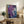 May include: Framed abstract art print with purple and blue swirling colors and white splatter details, displayed on rustic wooden console table alongside fern plant, books, ceramic vase, and brown leather chair in bright living room setting