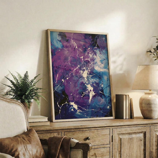 May include: Framed abstract art print with purple and blue swirling colors and white splatter details, displayed on rustic wooden console table alongside fern plant, books, ceramic vase, and brown leather chair in bright living room setting