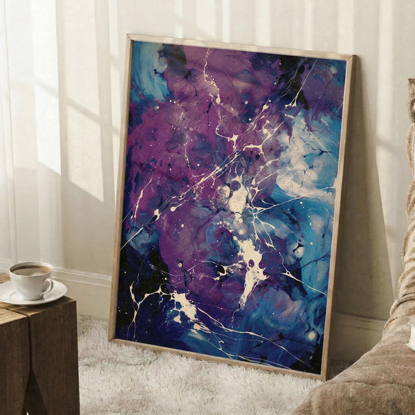 May include: Large framed abstract art print leaning against white wall corner, featuring vibrant purple and blue fluid paint splatters with white crackling details and scattered dots. Natural wood frame sits on textured white rug beside wooden side table with white coffee cup. Cozy room setting with beige blanket visible.