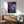 May include: Abstract artwork with purple, blue and teal fluid paint effects and golden crackling lines displayed in a thin wooden frame above a mid-century modern wood credenza. Scene shows sage green upholstered armchair with wooden arms, white ceramic table lamp, and natural sunlight casting shadows on white walls.