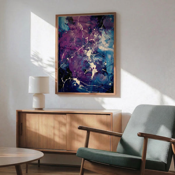 May include: Abstract artwork with purple, blue and teal fluid paint effects and golden crackling lines displayed in a thin wooden frame above a mid-century modern wood credenza. Scene shows sage green upholstered armchair with wooden arms, white ceramic table lamp, and natural sunlight casting shadows on white walls.