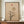 May include: Framed minimalist artwork displayed on wooden credenza featuring abstract tree silhouette with bare branches against beige textured background, orange sun, large tan circle, and geometric mountain shapes. White ceramic vase and stacked books flank the dark wood frame in neutral room setting with cream walls and white molding.