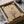 Minimalist artwork in black frame being unboxed from cardboard shipping box with protective wrapping. Features bare branched tree silhouette against beige background with geometric shapes - small red circle, larger tan circle, and curved forms suggesting abstract landscape. Textured paper surface visible with subtle linear details. Frame surrounded by white tissue paper and cardboard packaging materials.