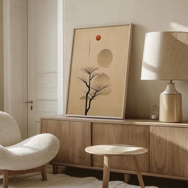 May include: Minimalist framed poster featuring bare tree silhouette with geometric beige circles and small red dot on cream background, displayed on wood sideboard next to ceramic table lamp in neutral-toned room with modern chair and white door