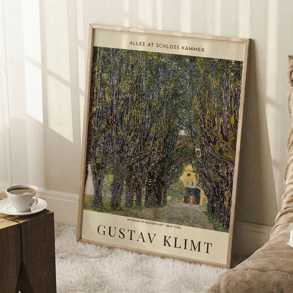 May include: Framed art print leaning against white wall beside beige chair and wooden side table with coffee cup. Print shows tree-lined pathway leading to yellow building, painted in impressionist style with dense green foliage creating natural archway. Cream mat border displays artist name Gustav Klimt and museum attribution. White fluffy rug beneath frame.