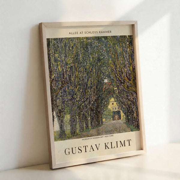 May include: Framed art print leaning against white wall showing tree-lined path leading to yellow building. Artwork features dense canopy of leafy trees creating tunnel effect in green and brown tones. Light wooden frame on cream matting. Museum attribution text visible at bottom. Natural lighting creates soft shadows on wall.