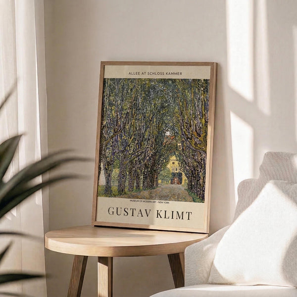 May include: Framed art print featuring a tree-lined avenue leading to a yellow building, displayed on a round wooden side table next to a beige upholstered chair. The poster shows Gustav Klimt's landscape painting with dense green foliage forming a natural tunnel over a path. Sunlight streams through white curtains creating soft shadows on neutral walls. A plant's leaves are visible in the corner, adding natural texture to the minimalist room setting.