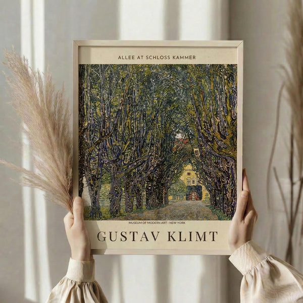 May include: Person in cream sweater holding framed art print featuring tree-lined path leading to yellow building. Artwork shows impressionistic style with dense green foliage creating natural archway. Light wood frame with beige matting displaying museum attribution text. Dried pampas grass visible to left, soft natural lighting from window creates gentle shadows on neutral background.