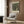 May include: Beige upholstered armchair with geometric patterned pillow positioned next to brass floor lamp with white circular base. Framed vintage art poster showing tree-lined pathway in green and yellow tones hangs on wood-paneled wall. Dark round side table with open book visible on left. Neutral tones throughout modern interior setting with natural lighting from window.
