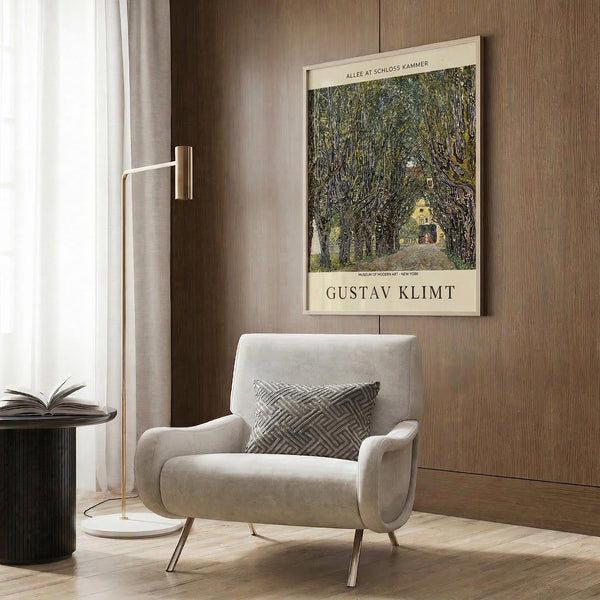 May include: Beige upholstered armchair with geometric patterned pillow positioned next to brass floor lamp with white circular base. Framed vintage art poster showing tree-lined pathway in green and yellow tones hangs on wood-paneled wall. Dark round side table with open book visible on left. Neutral tones throughout modern interior setting with natural lighting from window.