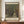 May include: Framed poster featuring Gustav Klimt's tree-lined pathway painting with dense green foliage forming a natural tunnel leading to a yellow building. Museum exhibition poster format with cream matting and artist name at bottom. Displayed on wooden cabinet beside white ceramic vase and stacked books in neutral-toned room with white paneled walls.