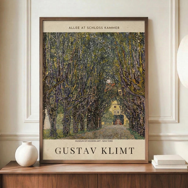 May include: Framed poster featuring Gustav Klimt's tree-lined pathway painting with dense green foliage forming a natural tunnel leading to a yellow building. Museum exhibition poster format with cream matting and artist name at bottom. Displayed on wooden cabinet beside white ceramic vase and stacked books in neutral-toned room with white paneled walls.