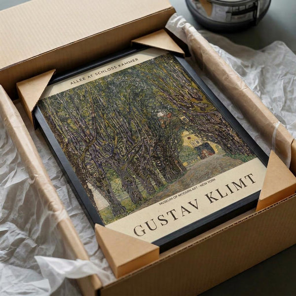 May include: Gustav Klimt art print in wooden frame being unpacked from cardboard box with tissue paper. The artwork shows a tree-lined path leading to a yellow building, painted in earthy greens and browns with Klimt's distinctive style. Museum of Modern Art New York attribution visible at bottom. Frame sits among white tissue paper with wooden corner protectors.