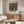 May include: Gustav Klimt museum poster featuring tree-lined path leading to yellow building, displayed above wooden sideboard in minimalist room with white curved chair, glass coffee table, and warm natural lighting creating reflections on surfaces.
