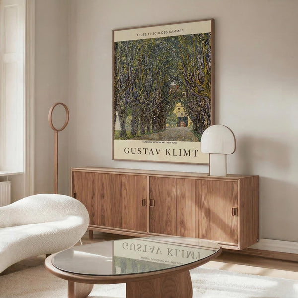 May include: Gustav Klimt museum poster featuring tree-lined path leading to yellow building, displayed above wooden sideboard in minimalist room with white curved chair, glass coffee table, and warm natural lighting creating reflections on surfaces.