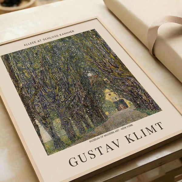 May include: Gustav Klimt art print displayed on white surface next to cream ribbon. The artwork shows a tree-lined avenue leading to yellow buildings, rendered in Klimt's distinctive style with intricate patterns and rich greens, blues, and earth tones. The print features white matting and text identifying it as a Museum of Modern Art New York piece.