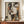 May include: Framed abstract portrait artwork in Art Deco style displayed on wooden credenza. The geometric composition features a stylized woman's face rendered in earthy tones of beige, olive green, terracotta, and brown with angular shapes and clean lines. Dark wood frame complements the mid-century modern setting with white ceramic vase and stacked books as styling elements.
