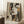 May include: Framed abstract art deco style portrait of a woman in earth tones including beige, olive green, rust orange, and brown geometric shapes. The stylized face features angular eyebrows, almond eyes, and defined lips in a cubist composition. The artwork leans against a white wall corner on a white textured rug, with a coffee cup on a wooden block nearby and part of a beige throw blanket visible.