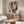 May include: Art deco style portrait of a woman rendered in geometric shapes and earth tones including sage green, rust orange, cream, and brown. The abstract face features angular forms, curved eyebrows, and stylized features. Displayed as a framed print above a mid-century modern walnut credenza with sliding doors, alongside a white sculptural chair, glass-top oval coffee table, and minimalist table lamp in a bright contemporary living room.