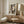 May include: Abstract art deco style portrait of a woman composed of geometric shapes in earth tones - beige, brown, orange, and green blocks forming facial features. White matted frame displayed on light wood credenza next to cream table lamp and small glass bottle. Modern mid-century room setting with textured cream chair, round wooden side table, white door, and neutral textured wallpaper.