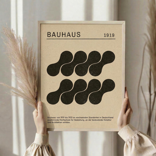 May include: Person holding framed Bauhaus poster with beige background and dark green geometric shapes arranged in two rows. Black text reads "BAUHAUS 1919" at top with German description below. Light wood frame, soft natural lighting, dried pampas grass visible on left side, neutral home decor setting.