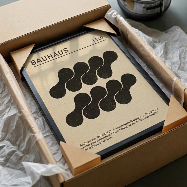 May include: A framed Bauhaus poster displayed in an unboxing scene with brown cardboard packaging and white tissue paper. The cream-colored print features black organic blob shapes arranged in a pattern, with "BAUHAUS" and "1919" text at the top and German text at the bottom. The black frame sits partially in the opened box alongside wooden dowels used for packaging protection.