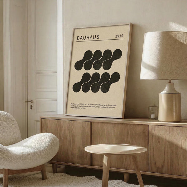May include: Framed Bauhaus poster with black geometric circles arranged in two rows on cream background, displayed on wooden sideboard next to ceramic table lamp in minimalist living room with neutral tones, white chair, and natural lighting creating soft shadows on textured walls.