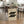 May include: Cream-colored poster with black circular geometric pattern leaning against wooden dining table in modern kitchen setting. Design features "BAUHAUS 1919" text at top with two rows of overlapping black circles below and German text at bottom. Poster shown in bright, minimalist dining area with wicker chairs, spider plant, wine bottle, and white cabinets visible in background.
