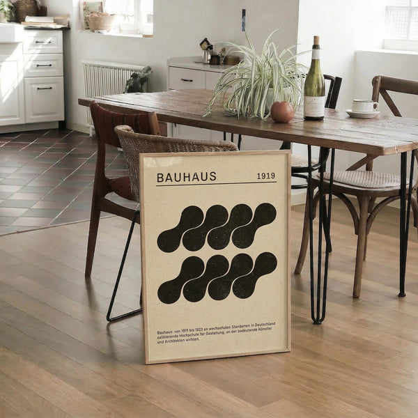 May include: Cream-colored poster with black circular geometric pattern leaning against wooden dining table in modern kitchen setting. Design features "BAUHAUS 1919" text at top with two rows of overlapping black circles below and German text at bottom. Poster shown in bright, minimalist dining area with wicker chairs, spider plant, wine bottle, and white cabinets visible in background.