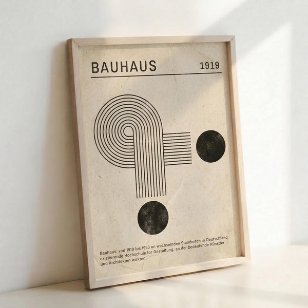 Framed geometric poster featuring "BAUHAUS 1919" text at top, with abstract line art forming curved and straight patterns, two dark circular shapes, all in black on cream background. Light wood frame leans against white wall, showing German text at bottom describing the design school's history from 1919-1933.