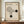 May include: Framed Bauhaus-inspired poster with beige textured background, featuring black geometric shapes - curved lines forming an abstract design and two solid circles. German text below describes the Bauhaus movement from 1919-1933. Displayed on wooden sideboard next to white ceramic vase and stacked books against cream paneled wall.