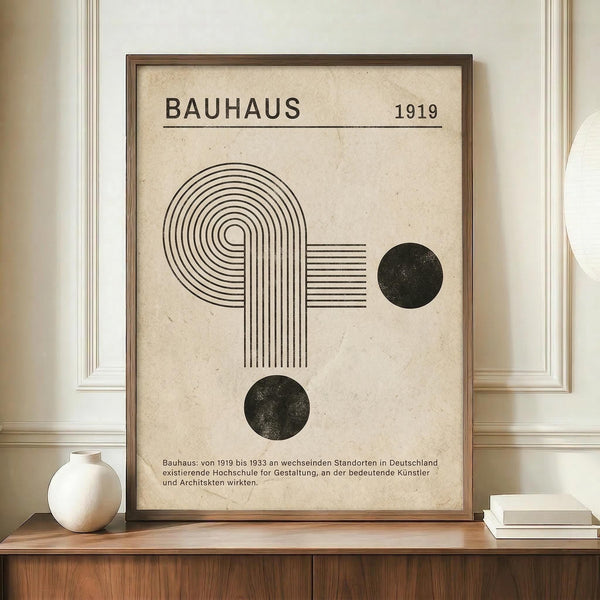 May include: Framed Bauhaus-inspired poster with beige textured background, featuring black geometric shapes - curved lines forming an abstract design and two solid circles. German text below describes the Bauhaus movement from 1919-1933. Displayed on wooden sideboard next to white ceramic vase and stacked books against cream paneled wall.