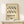 May include: Framed poster with wavy black line pattern and "Bauhaus" text in German, displayed on white wooden dresser next to small wrapped gift with evergreen sprig. Natural wood frame against cream wall with soft lighting creating shadows.