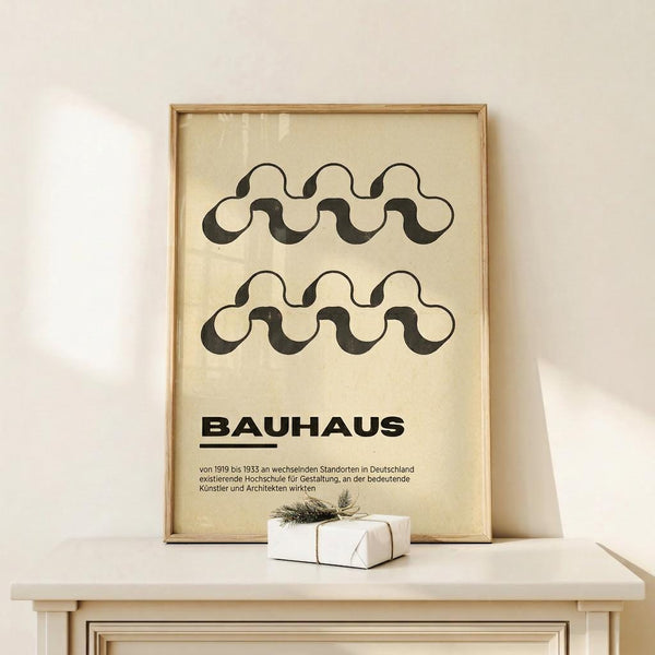 May include: Framed poster with wavy black line pattern and "Bauhaus" text in German, displayed on white wooden dresser next to small wrapped gift with evergreen sprig. Natural wood frame against cream wall with soft lighting creating shadows.