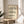 May include: Framed poster with cream background featuring two rows of black wavy interlocking patterns above bold "BAUHAUS" text and German description, displayed on light wood round stool next to beige upholstered chair and plant in bright sunlit room with white walls and curtains