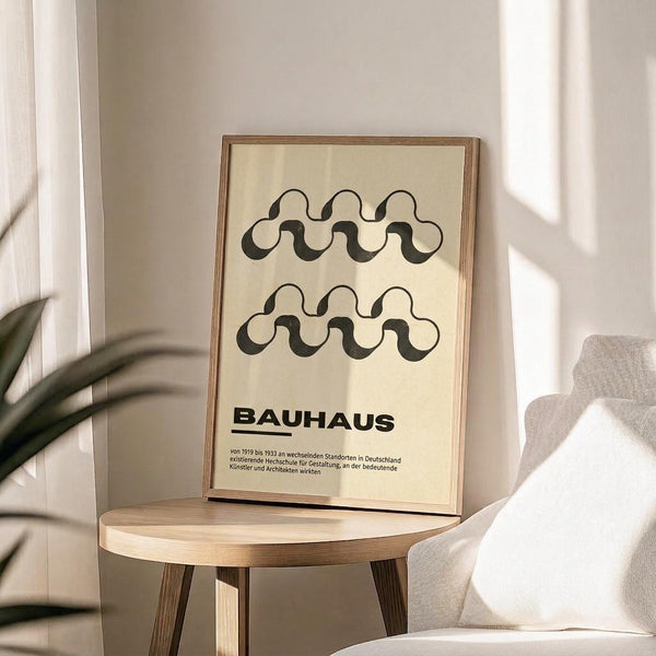 May include: Framed poster with cream background featuring two rows of black wavy interlocking patterns above bold "BAUHAUS" text and German description, displayed on light wood round stool next to beige upholstered chair and plant in bright sunlit room with white walls and curtains