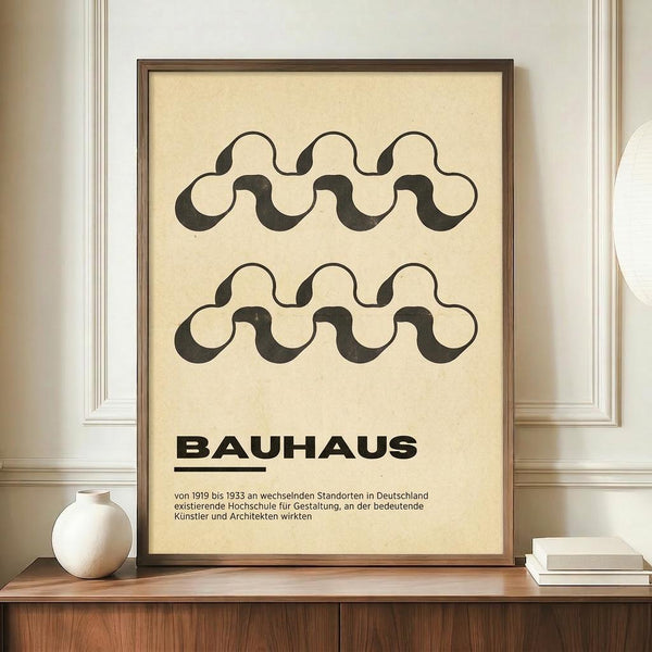 May include: Framed beige poster with black wavy geometric pattern and "BAUHAUS" text, displayed on wooden sideboard next to white ceramic vase and stacked books against cream paneled wall in modern interior setting