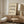 May include: Framed Bauhaus poster with black wavy geometric patterns on cream background, displayed on wooden sideboard next to beige ceramic table lamp in minimalist neutral-toned room with white upholstered chair and small wooden stool.