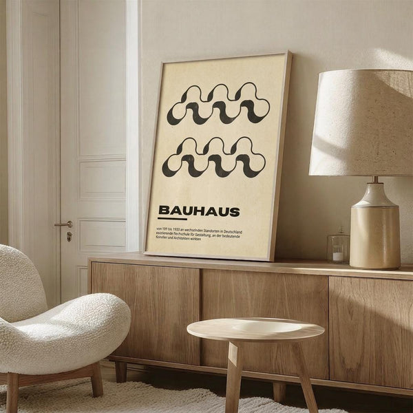 May include: Framed Bauhaus poster with black wavy geometric patterns on cream background, displayed on wooden sideboard next to beige ceramic table lamp in minimalist neutral-toned room with white upholstered chair and small wooden stool.
