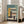 May include: Framed Bauhaus-inspired architectural poster showing geometric building design with yellow, teal, coral and cream colored blocks, displayed on wooden floor next to beige upholstered chair in bright room with white walls and windows.