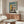 May include: Bauhaus-inspired architectural poster with geometric building design in teal, orange, and cream colors, displayed in wooden frame above mid-century modern walnut credenza. Room features white sculptural chair, glass oval coffee table, and minimalist white table lamp, creating clean Scandinavian interior styling.