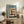 May include: Framed architectural poster with geometric Bauhaus-style building illustration in muted teal, mustard yellow, cream and coral colors, displayed on rustic wooden sideboard alongside fern plant, books, ceramic vase and leather armchair in cozy living room setting with white walls and natural lighting.
