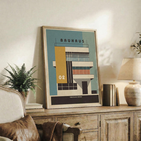 May include: Framed architectural poster with geometric Bauhaus-style building illustration in muted teal, mustard yellow, cream and coral colors, displayed on rustic wooden sideboard alongside fern plant, books, ceramic vase and leather armchair in cozy living room setting with white walls and natural lighting.