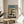 May include: Framed modernist architectural poster featuring a geometric building design in teal, yellow, coral, and cream colors with "BAUHAUS" text and "02" number. The wooden-framed print sits on a round light wood side table next to a beige upholstered chair, with plant leaves and natural sunlight creating shadows on white walls in a minimalist interior setting.