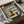 May include: Framed Bauhaus-style architectural poster in cardboard shipping box with protective tissue paper. The modernist print features geometric building design in sage green, mustard yellow, coral, and cream colors with "BAUHAUS" and "02" text. Black frame sits in brown cardboard packaging on white surface next to wooden dowel and tape dispenser.