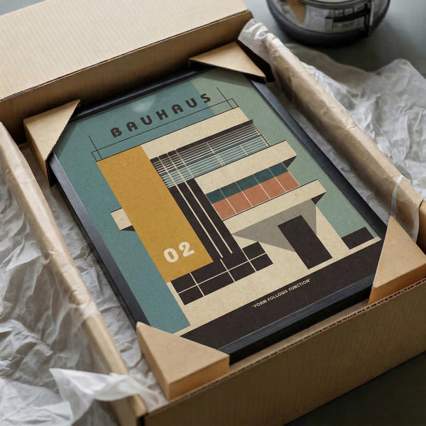 May include: Framed Bauhaus-style architectural poster in cardboard shipping box with protective tissue paper. The modernist print features geometric building design in sage green, mustard yellow, coral, and cream colors with "BAUHAUS" and "02" text. Black frame sits in brown cardboard packaging on white surface next to wooden dowel and tape dispenser.