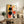 May include: Abstract geometric poster with black circles, orange and red shapes, and colorful rectangles in light wood frame, displayed on rustic wooden sideboard alongside fern plant, books, ceramic lamp, and brown leather chair in warm-toned living room setting.