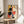 May include: Framed abstract geometric poster with colorful circles, rectangles, and lines in Bauhaus style, displayed on light wooden round side table next to beige upholstered chair. Artwork features black circles, orange and red shapes, blue rectangles, and yellow sections against cream background in natural wood frame. Soft natural lighting creates window shadows on white wall, with plant leaves visible in corner.