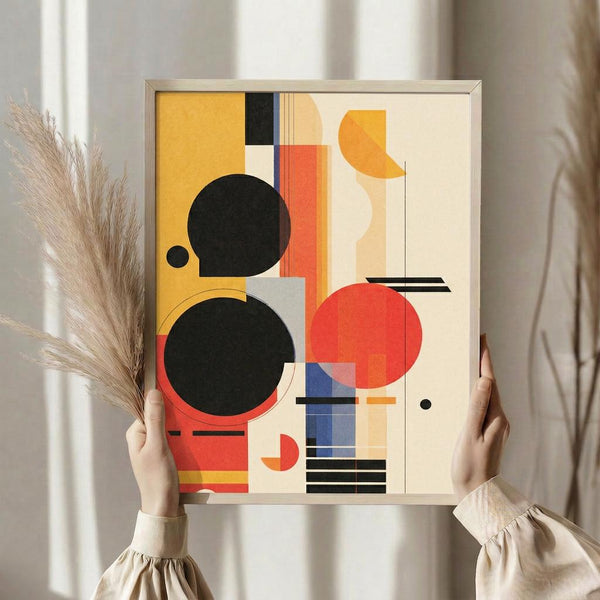 May include: Person holding a framed abstract geometric art print featuring bold circles, rectangles, and lines in black, red, orange, yellow, and blue on cream background. Modern Bauhaus-style composition with overlapping shapes and clean geometric forms. Silver frame, neutral room setting with dried pampas grass and beige curtains visible.